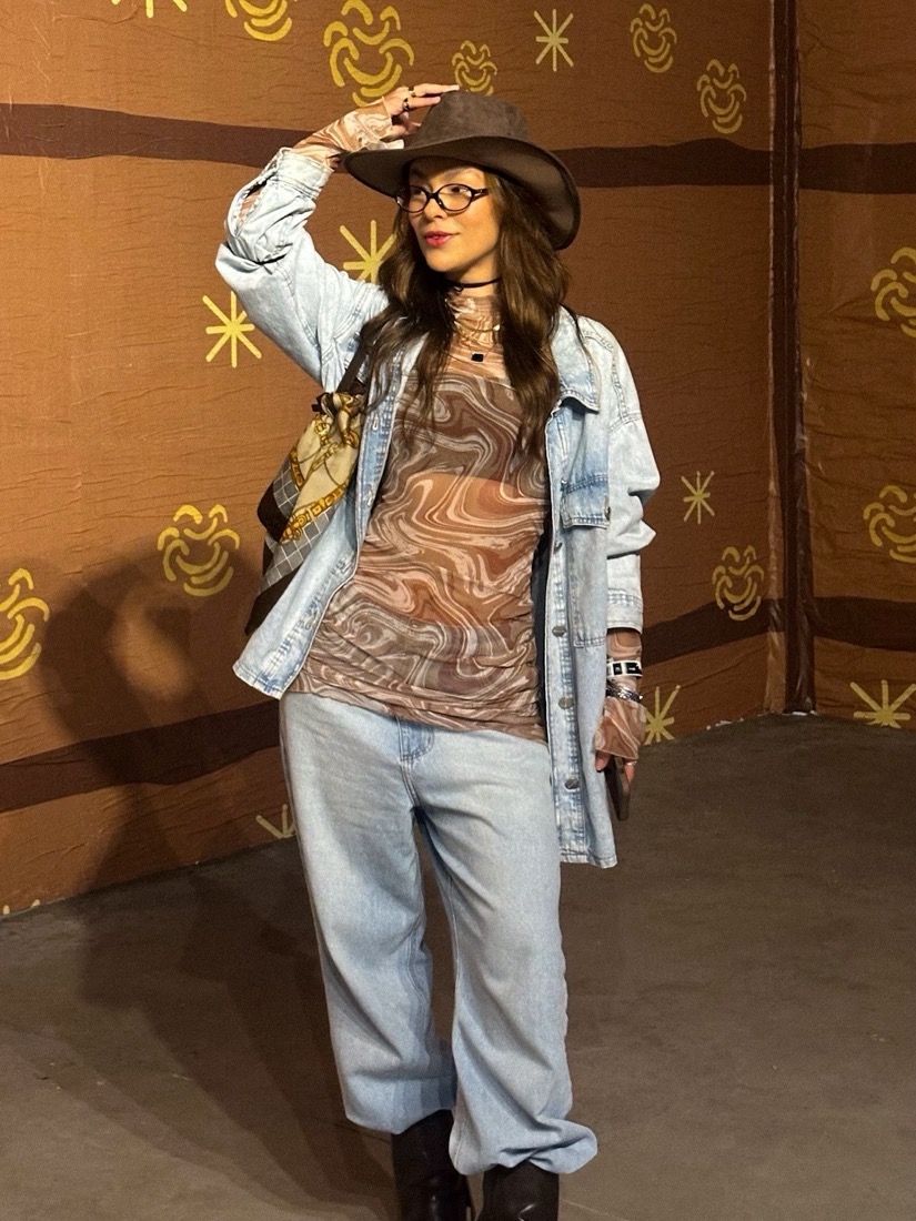 Carol de chapéu marrom, camisa e calça jeans claras e blusa estampada